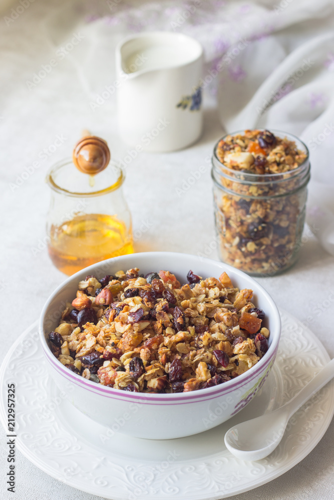 Breakfast: fresh oat granola in bowl with nuts, dried fruits, seeds and honey with milk in a on light background.