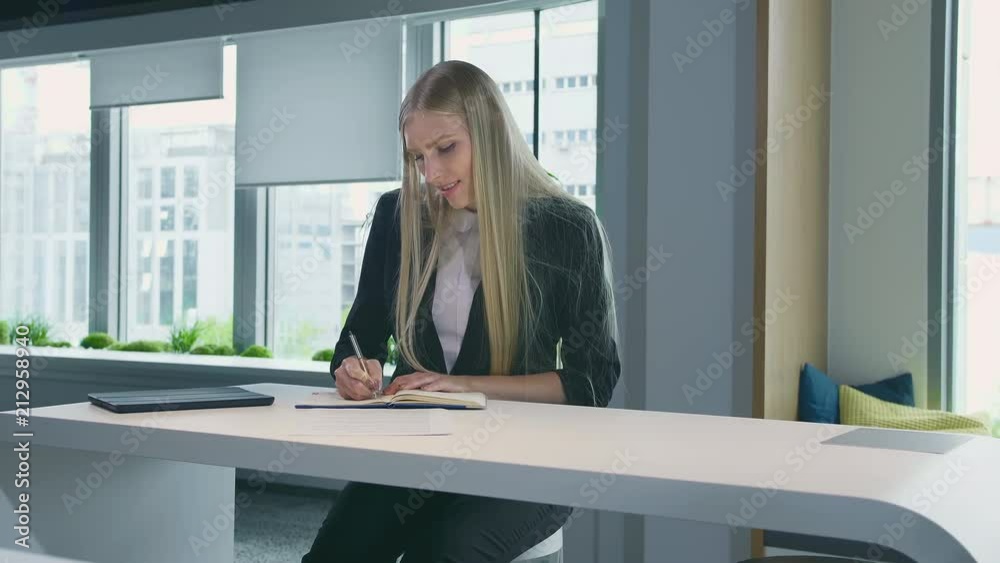 Elegant woman working in stylish office. Modern blond woman in trendy suit sitting at table in light contemporary office and writing on papers. Elegant woman working in stylish office. Modern blond woman in trendy suit sitting at table in light contemporary office and writing on papers.