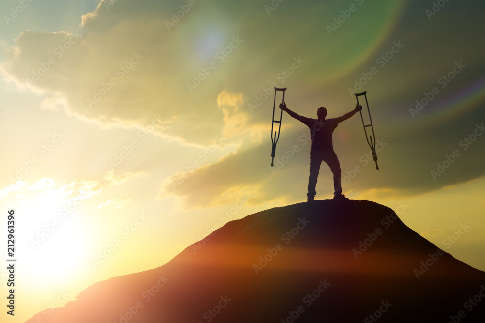 silhouette of a happy man on top of a mountain with crutches, lifting her  arms up, facing dramatic sky at dawn. The cure of the disease,  rehabilitation, healing, trauma, medicine. Photos