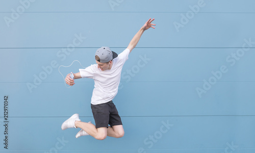 Young man in a white T-shirt and summer clothes listens to music in headphones and jumps on a blue background.Man jumping on the background of a blue wall with headphones and a smartphone in his hands