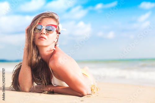 young longhaired woman in sunglasses and bikini smiling and sunbathing by the beach photo