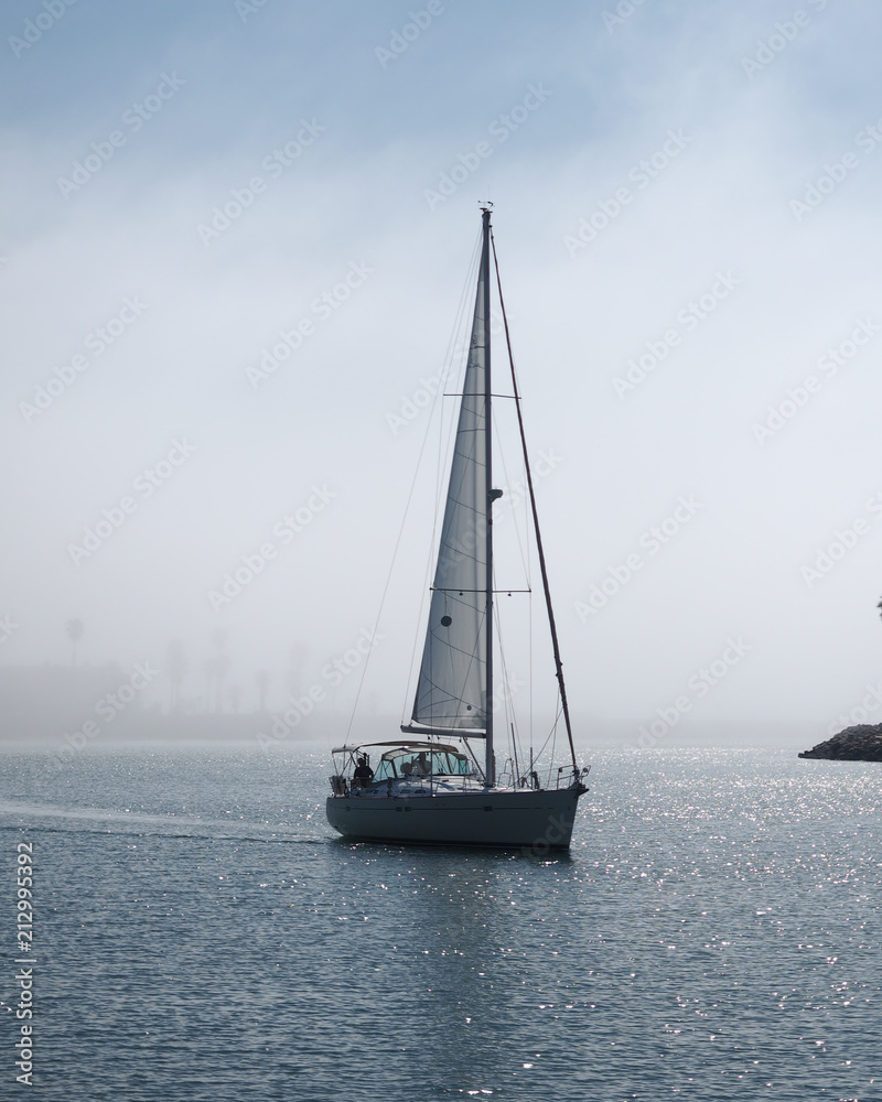 A sailboat in the water with palm trees seen through the foggy background and blue skies with clouds