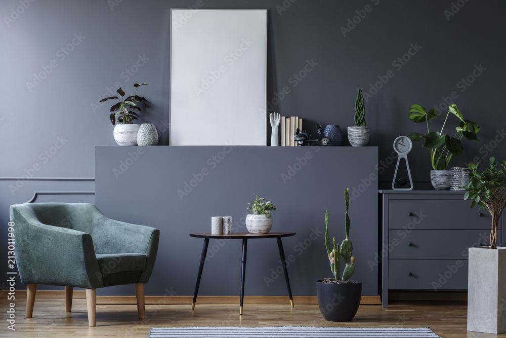 Green armchair next to table and plant in grey living room interior with mockup of poster. Real photo