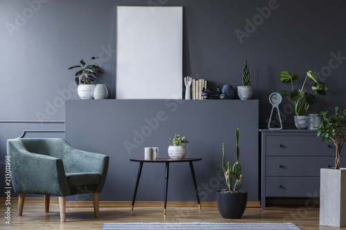 Green armchair next to table and plant in grey living room interior with mockup of poster. Real photo