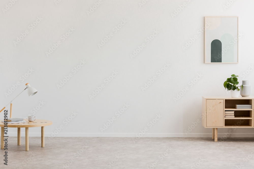 Desk lamp on a small table and a simple, wooden cabinet in an empty living room interior with white wall and place for a sofa. Real photo. - obrazy, fototapety, plakaty 
