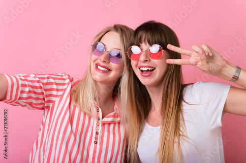 Two young girl friends standing together and having fun. Showing signs with their hands. Looking at camera and smiling. Inside