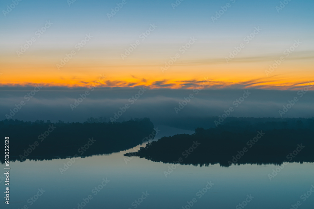 Early blue sky reflected in river water. Riverbank with forest under predawn sky. Yellow stripe in picturesque sky. Fog hid trees on island. Colorful morning atmospheric landscape of majestic nature.