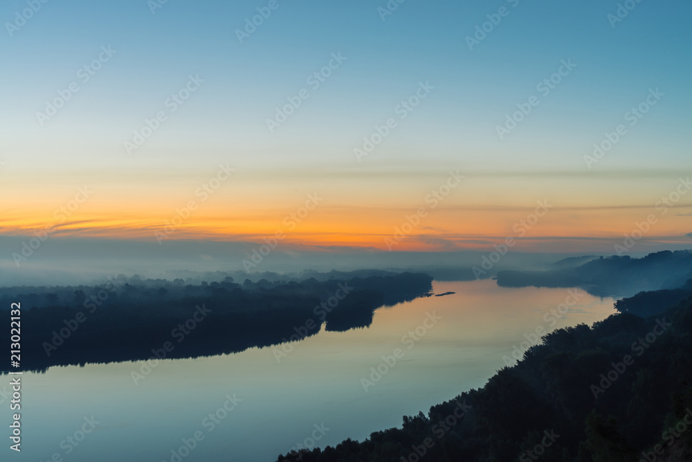 View from high shore on river. Riverbank with forest under thick fog. Dawn reflected in water. Yellow stripe in picturesque predawn sky. Mystical calm morning atmospheric landscape of majestic nature.