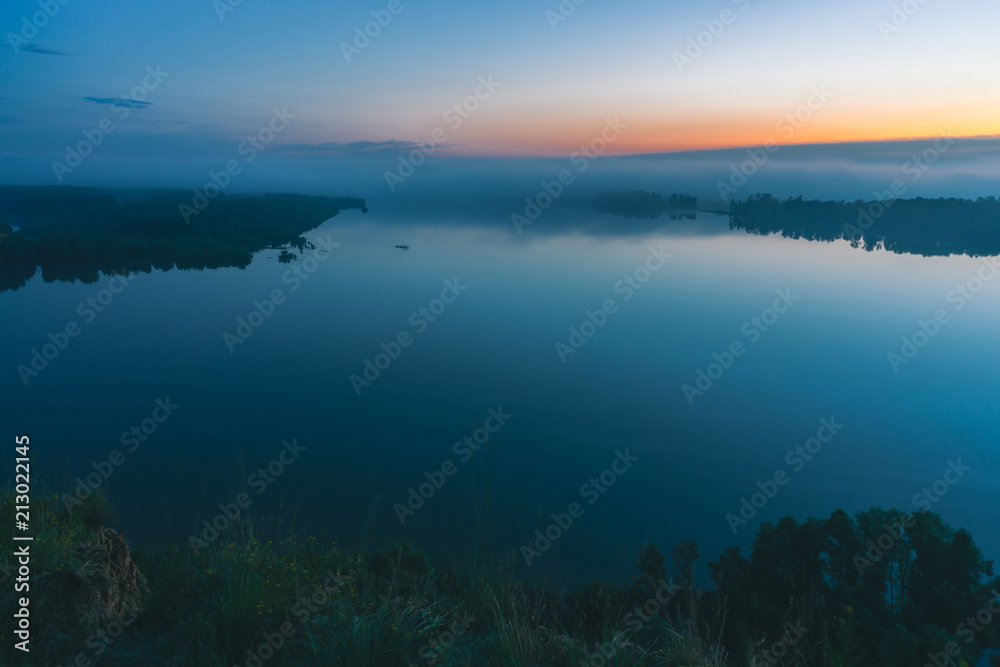 Mystical view from high shore on broad river. Riverbank with forest under mist. Early haze above trees. Orange glow in picturesque predawn sky. Morning atmospheric calm landscape of majestic nature.