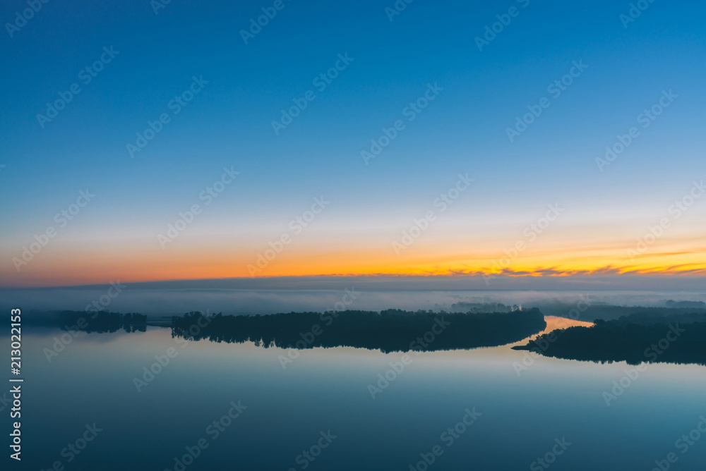 Broad river flows along diagonal shore with forest under thick fog. Early blue sky reflected in water. Yellow glow in picturesque predawn sky. Colorful morning atmospheric landscape of majestic nature