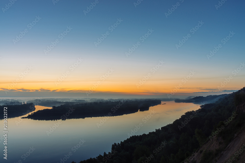 View from high shore on broad river. Riverbank with forest under thick fog. Dawn reflected in water. Yellow glow in picturesque predawn sky. Colorful morning atmospheric landscape of majestic nature.