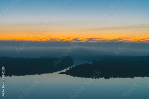 Early blue sky reflected in river water. Riverbank with forest under predawn sky. Yellow stripe in picturesque sky. Fog hid trees on island. Colorful morning atmospheric landscape of majestic nature.