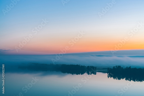 Broad mystical river flows along diagonal shore with silhouette of trees and thick fog. Orange and pink glow in predawn vanila sky. Morning atmospheric landscape of majestic nature in tender tones. © Daniil