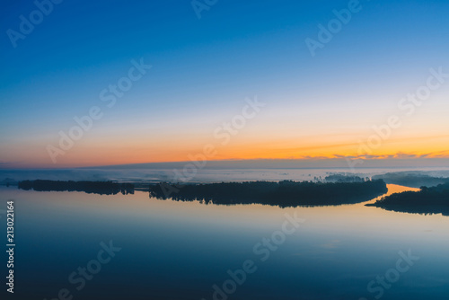Broad river flows along diagonal shore with forest under thick fog. Early blue sky reflected in water. Yellow glow in picturesque predawn sky. Colorful morning atmospheric landscape of majestic nature