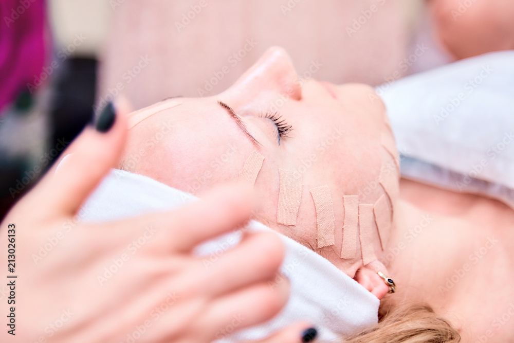 Fototapeta premium Young woman lying with tape on her face. Hands of the cosmetologist. Kinesiotaping. Physiotherapy and cosmetology procedure. Method of non-surgical skin rejuvenation. Facial skin care.