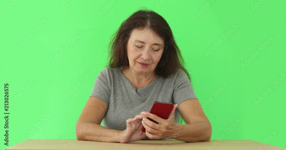 Mature casual woman with long brown hair sitting at table and surfing smartphone on green chromakey background Mature casual woman with long brown hair sitting at table and surfing smartphone on green chromakey background