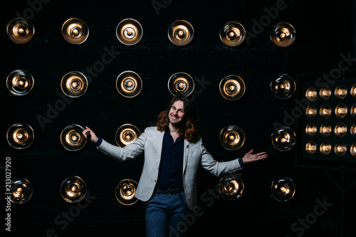 a man in a white jacket and in a black shirt, with long hair, stands against the backdrop of bright lanterns, safites. on the stage.performance. smiles
