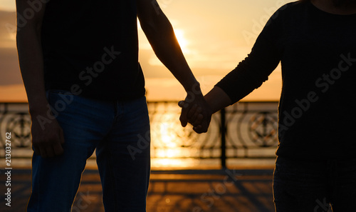 The loving couple is holding hands at sunset. The tall slender man and the low full girl are against the background of setting a sun.