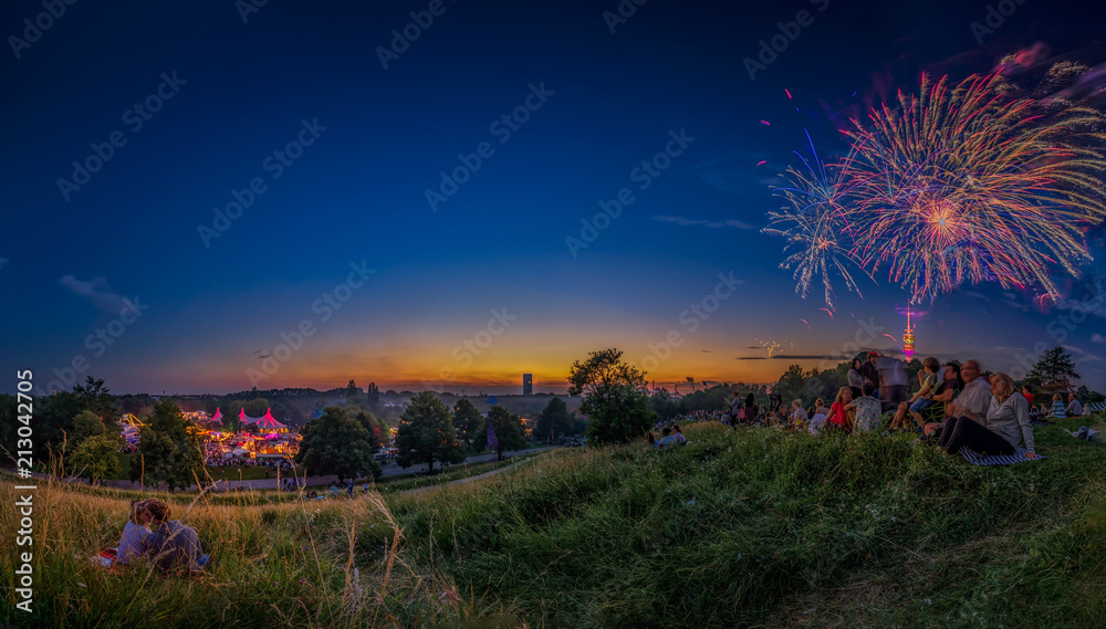 Fototapeta premium Das spektakuläre Feuerwerk im Olympiapark von München mit angrenzendem Tollwood Festival als Panorama am Abend bei herrlicher Stimmung zur blauen Stunde, Menschen haben Spaß und Freude an den Raketen