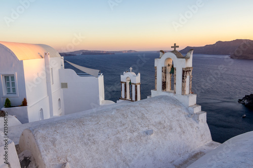 Church white roof, crosses and bells in Santorini, Greece. Amazing daytime sunset view towards the deep sea crystal waters with the caldera in the background