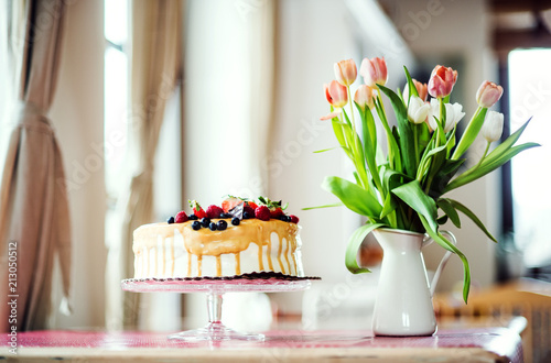 A birthday cake on a glass stand and a vase with tulips on the table.
