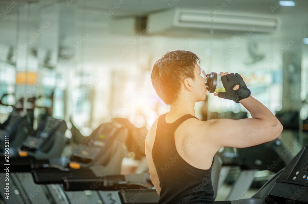 The sports young man drinks protein in a shaker in a gym. Sports concept.Handsome attractive muscular man after training in the gym.Young man drinking protein shake on treadmill after doing exercises ...