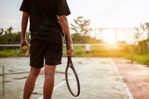 Close up of man holding tennis racket on clay court. In his hand is tennis ball. On court is sunset. © Day Of Victory Stu.
