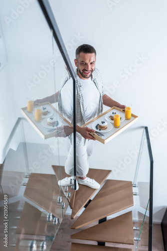 smiling man with breakfast on wooden tray walking up stairs at home