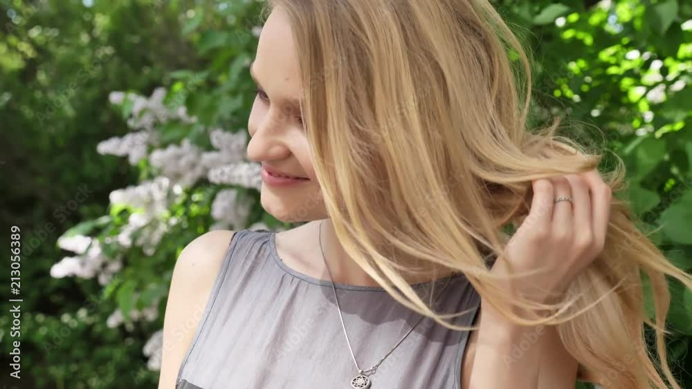 Young cute blond girl is watching at camera in park in daytime in summer, has her hair down, smiling Young cute blond girl is watching at camera in park in daytime in summer, has her hair down, smiling