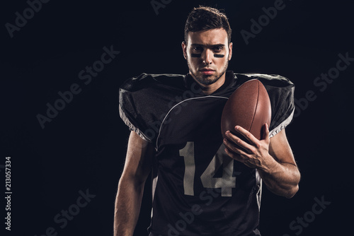 young serious american football player with ball looking at camera isolated on black