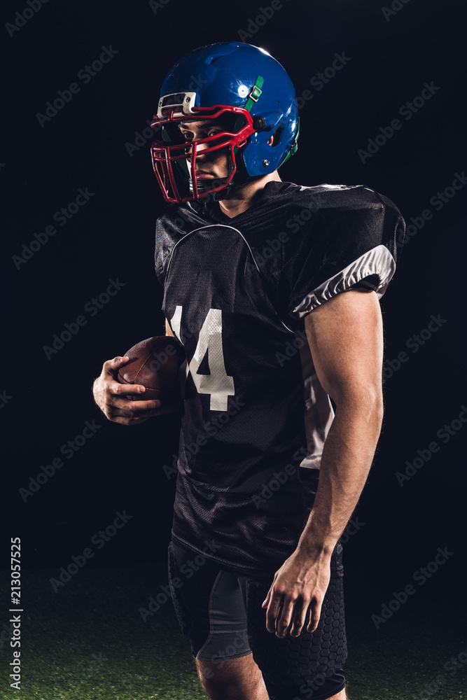 confident handsome american football player with ball looking away on black