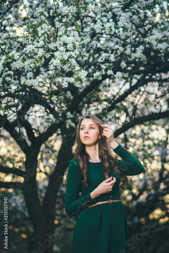 Young beautiful woman in green dress posing in the blooming garden at the sunset