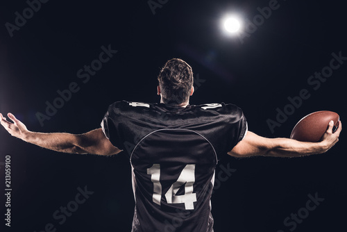 rear view of american football player with ball looking up with outstretched hands under spotlight on black