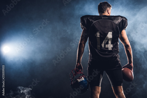 Fototapeta Naklejka Na Ścianę i Meble -  rear view of equipped american football player with helmet and ball in hands against white smoke