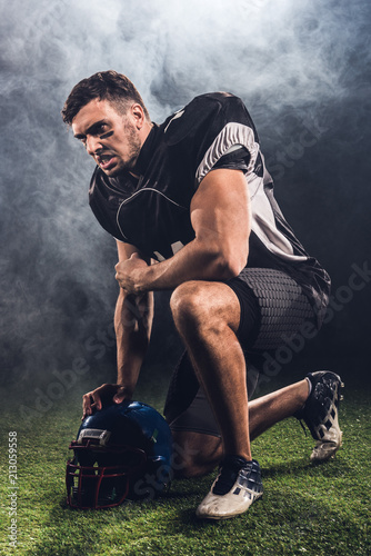 Fototapeta Naklejka Na Ścianę i Meble -  angry young american football player with helmet standing on knee on black