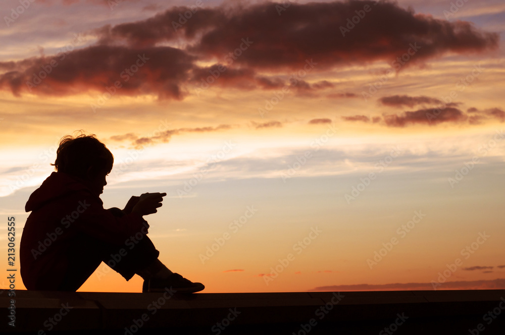 Lonely sad boy sits on the roof with phone in hands on sunset background