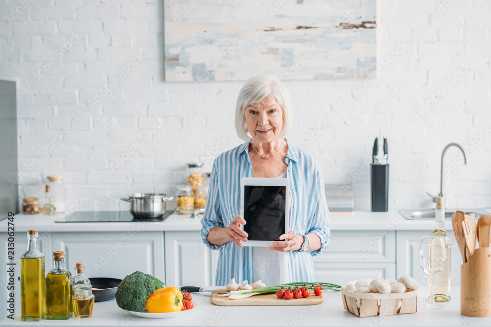 portrait of senior woman showing tablet with blank screen while standing at counter with fresh vegetables in kitchen portrait of senior woman showing tablet with blank screen while standing at counter with fresh vegetables in kitchen