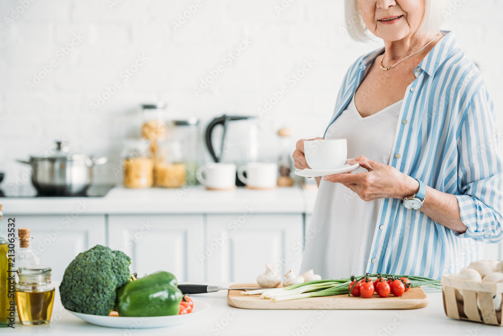 cropped shot of senior woman with cup of coffee standing at counter with fresh vegetables for dinner in kitchen