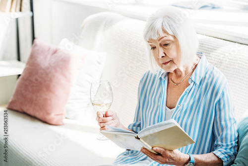 focused senior woman with glass of wine reading book on couch at home