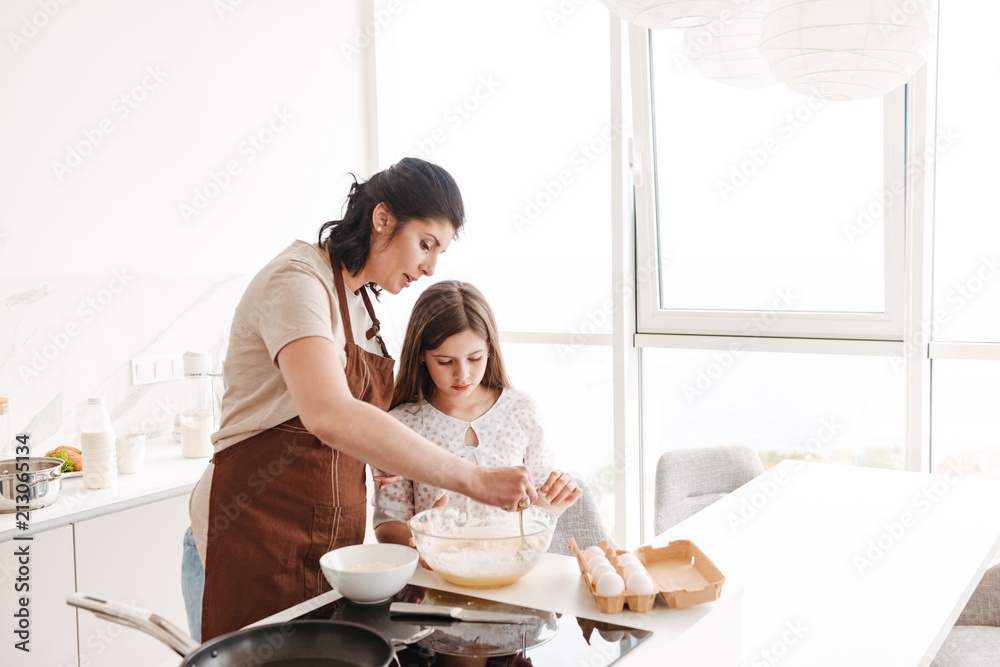 Satisfied content woman and little daughter baking pastry in kitchen at home, and kneading dough with spoon