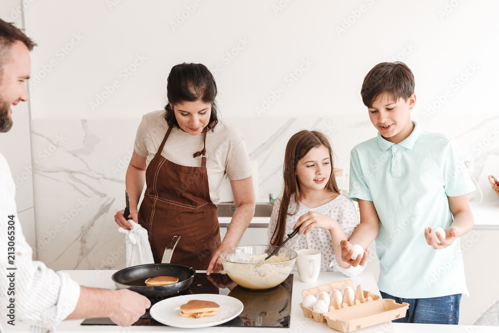 Caucasian family woman and man with happy kids boy and girl 8-10 cooking together, and frying pancakes on modern stove in kitchen Caucasian family woman and man with happy kids boy and girl 8-10 cooking together, and frying pancakes on modern stove in kitchen