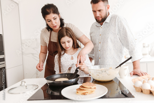 Happy parents woman and man with little daughter 8-10 cooking together, and frying pancakes on modern stove in kitchen at home