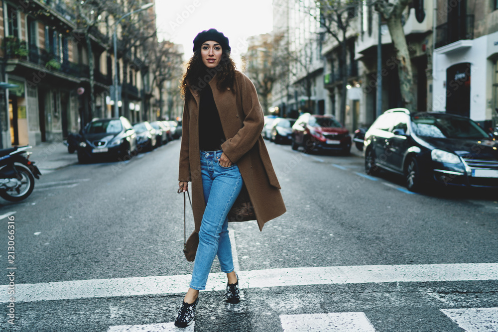 Fototapeta premium Elegant business woman enjoying walking the city after hard working day. Student girl in a trendy clothes crossing the road. Stylish hipster girl wearing oversized coat looking at the camera.
