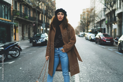 Beautiful model look woman with long dark curly hair wearing stylish clothes standing in the middle of the road on a blurred street background. Stylish blogger walking the city in the evening.