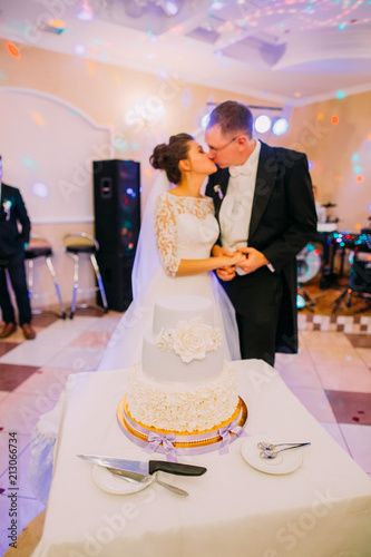 The white floral wedding cake at the background of the kissing newlywed couple. photo
