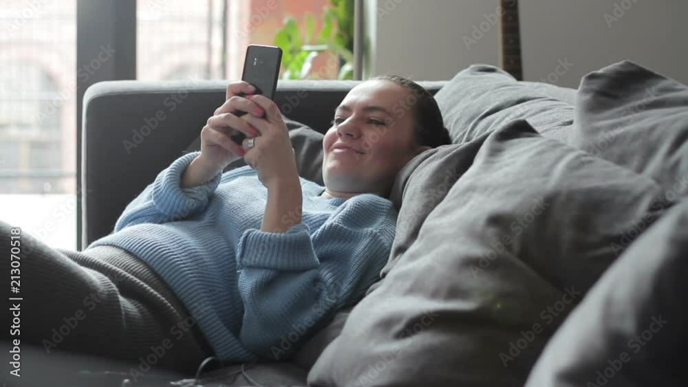 Smiling pretty woman reading a text on her smartphone while sitting on cosy sofa Smiling pretty woman reading a text on her smartphone while sitting on cosy sofa