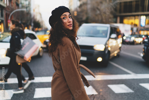 Portrait of a charming caucasian woman with beautiful long dark curly hair wearing coat and a beret looking back at the camera while crossing the street on a blurred urban background. © JKstock