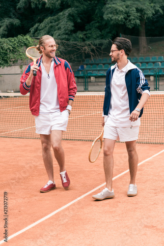 cheerful sportive friends with wooden rackets walking on tennis court