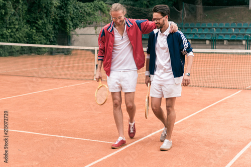 cheerful sportive friends with wooden rackets hugging and walking on tennis court