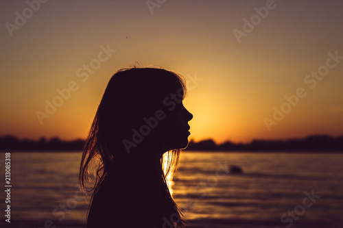Silhouette of young woman sitting near the water on sand beach with sand in hands © Andrei